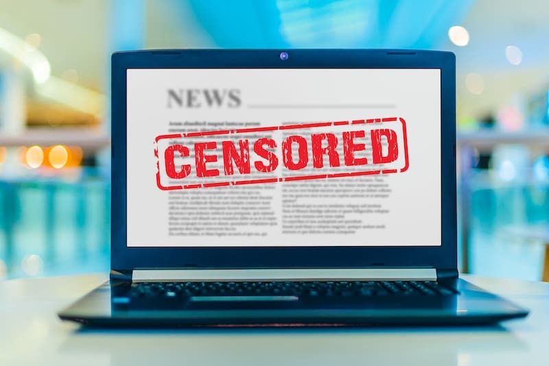 A laptop screen displays a news article with the word "CENSORED" stamped in bold red letters across the text, symbolizing restricted or suppressed information.