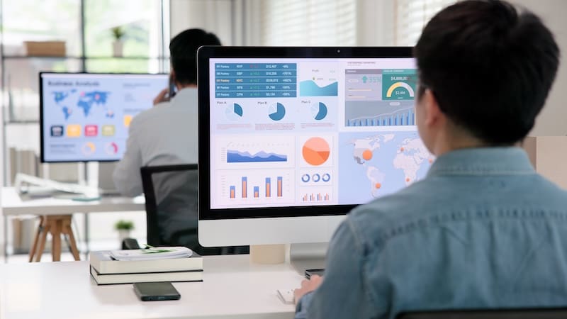 Two people sit at their desks on computers while looking at stats on the screen.