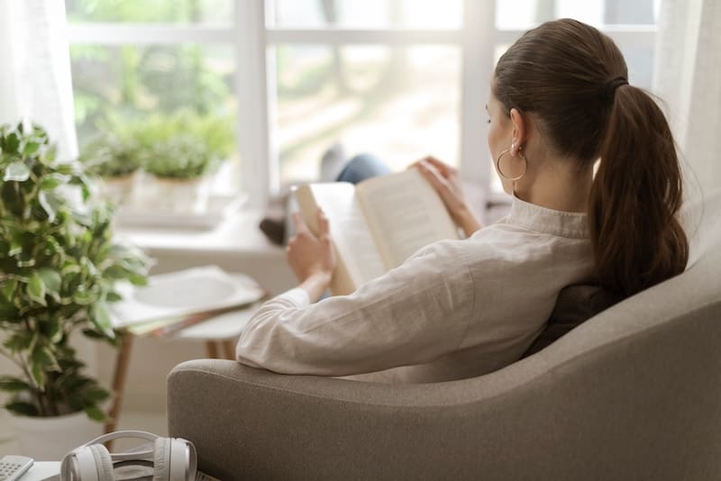 Young woman relaxing at home next to a window, she is sitting on the armchair and reading a book.