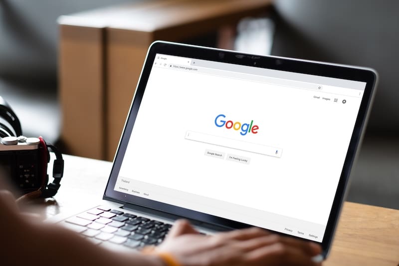 A person using a laptop with the Google search homepage open on the screen, sitting at a table with a camera and blurred background.