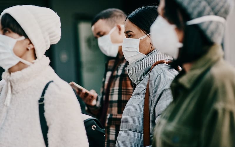 Four people wearing face masks and winter clothing stand close together outdoors; one person is looking at their phone, while the others look ahead.