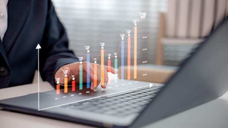 Businessman using laptop with rising monthly ecommerce sales chart and shopping cart icons, representing revenue growth, digital business analytics, and online retail strategy.