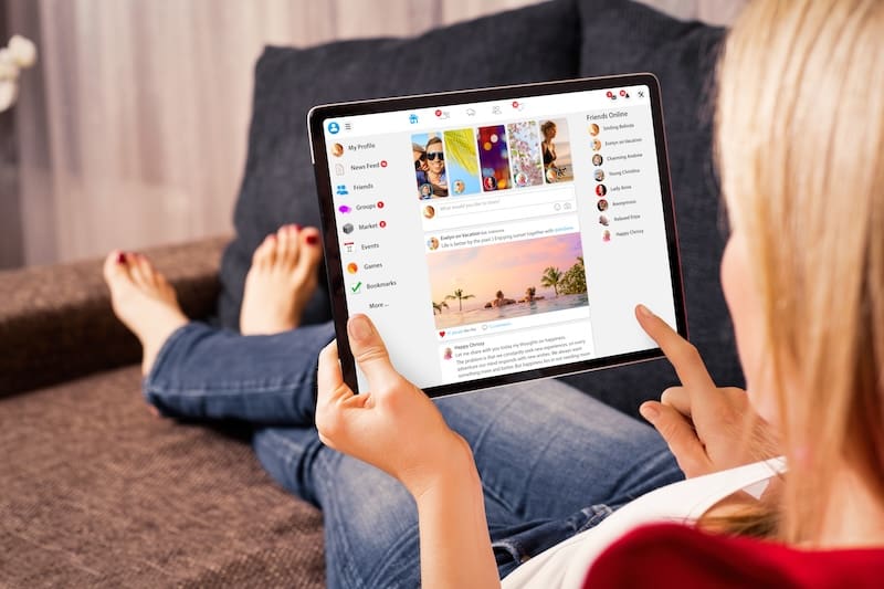 A person sits on a couch using a tablet, browsing a social media platform with photos and a list of friends online. The screen displays a beach sunset photo and profile icons.