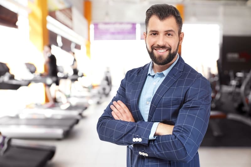 A smiling man with a beard and neatly styled hair, wearing a blue plaid suit jacket and light blue shirt, stands with arms crossed inside a modern gym, ready to offer smart replies to rude comments examples if needed.