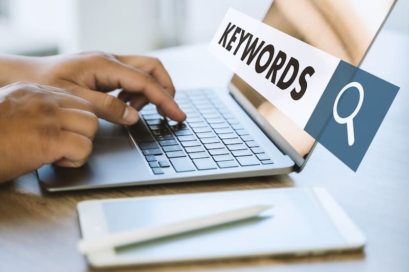 A person types on a laptop keyboard, with a transparent search bar graphic displaying the word “KEYWORDS” and a magnifying glass icon. A smartphone rests on the table nearby.
