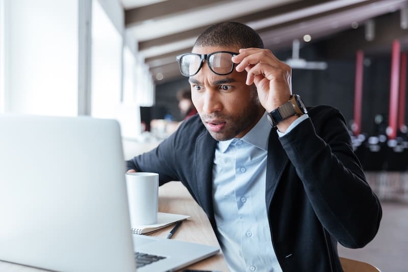 A man in a suit sitting at a desk looks surprised as he lifts his glasses while staring at a laptop screen, with a cup and papers nearby in a modern office setting.