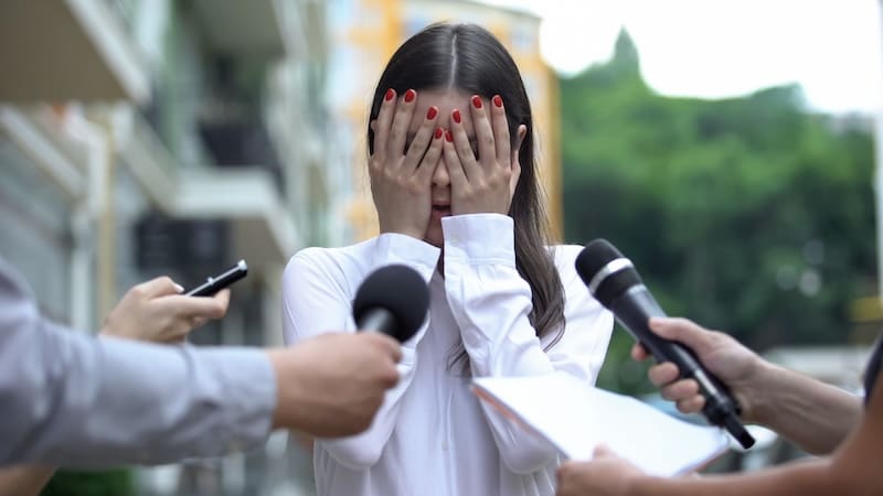 A woman in a white shirt covers her face with her hands while several microphones and a smartphone are held out toward her, suggesting she is overwhelmed by media attention.
