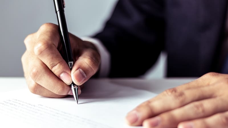 A person in a suit writes on a document with a black pen, with only their hands and part of their suit visible, suggesting a formal or business setting.