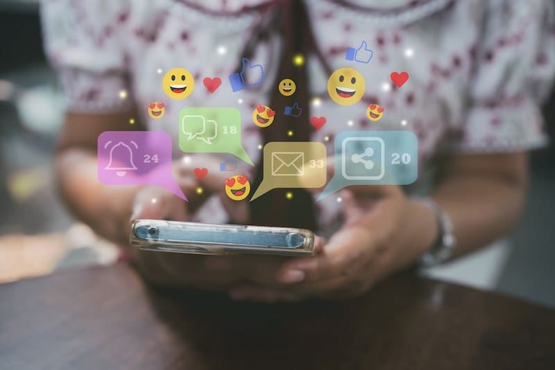 A person holding a smartphone with social media notification icons, such as likes, comments, and messages, floating above the screen. The person is seated at a table and wearing a patterned shirt.