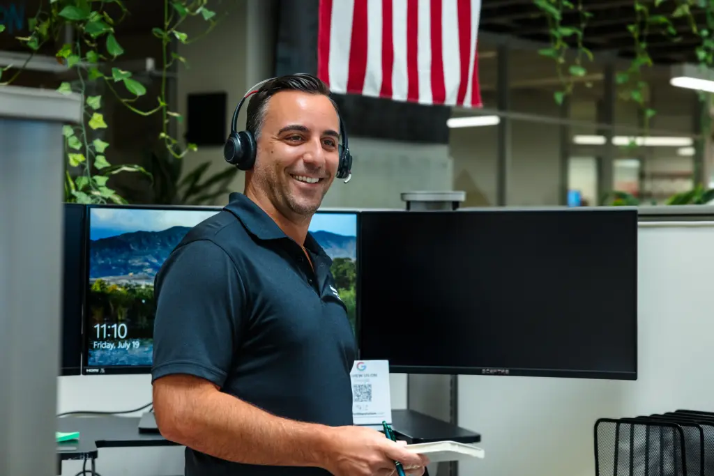 A man wearing a headset and a dark polo shirt smiles while standing near a computer desk in an office. An American flag hangs above, and a monitor displays a scenic wallpaper and the time 11:10.