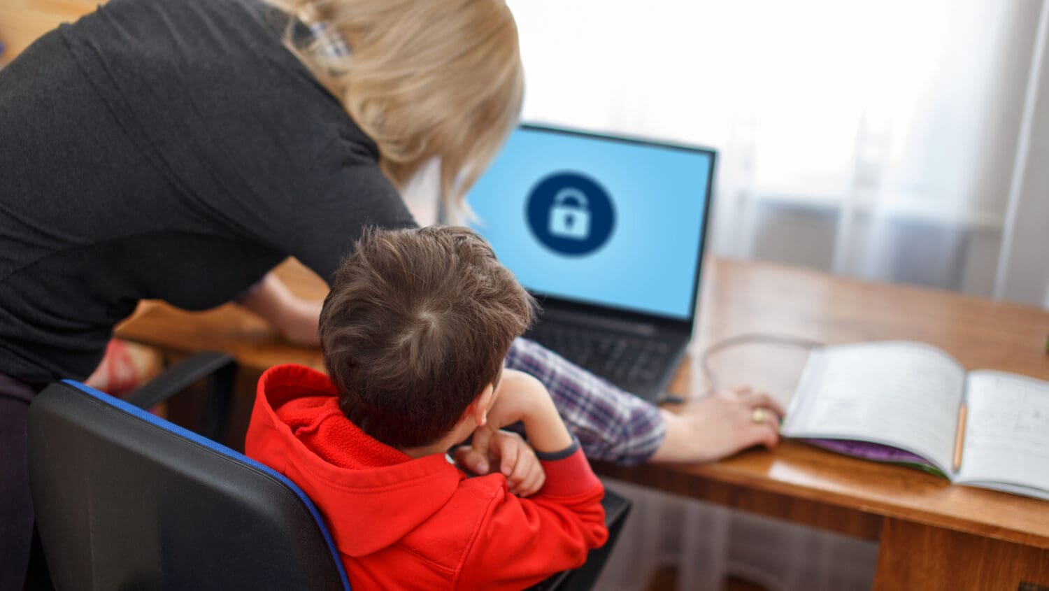 An adult helps a child sitting at a desk with a laptop displaying a padlock icon on the screen. An open notebook lies nearby, suggesting a learning or online safety scenario.
