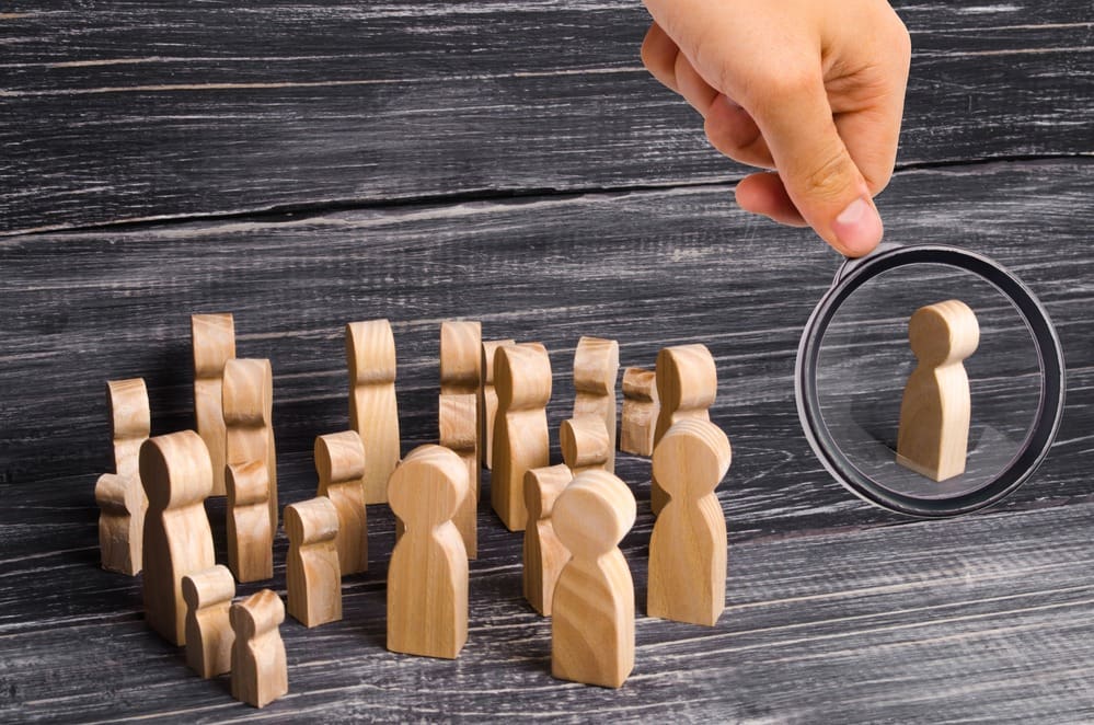 A hand is holding a magnifying glass over a group of wooden figures.