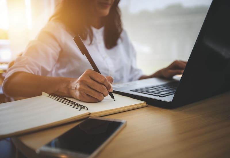 A woman diligently jotting down notes in her notebook, multitasking with a laptop on her desk.