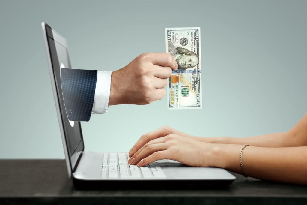 A woman is holding a dollar bill in front of a laptop.