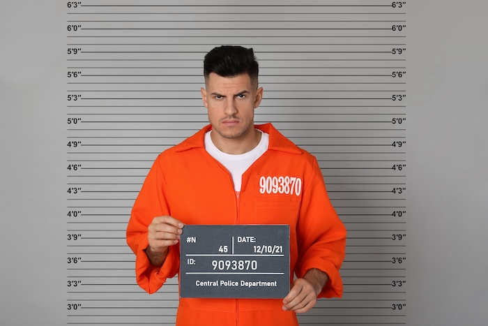 A man in an orange prison uniform holding up a mugshot, wondering how to get it removed.