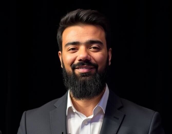 A bearded man in a dark suit and white shirt smiles confidently, embodying a professional team spirit as he stands against a black background.