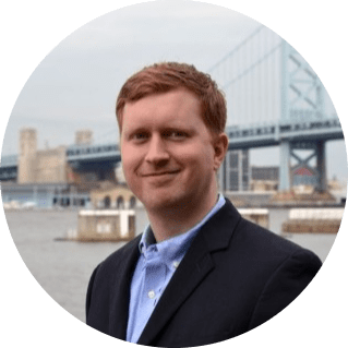 A man with short red hair, wearing a dark blazer and blue shirt, stands in front of a waterfront with a large suspension bridge and city buildings in the background on a cloudy day, ready to lead his team.