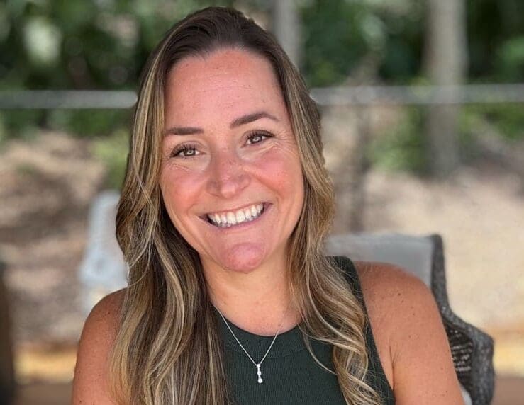 A woman with long, wavy light brown hair and a sleeveless dark green top smiles while sitting outdoors. She wears a silver necklace and a smartwatch, with greenery and a fence in the background.