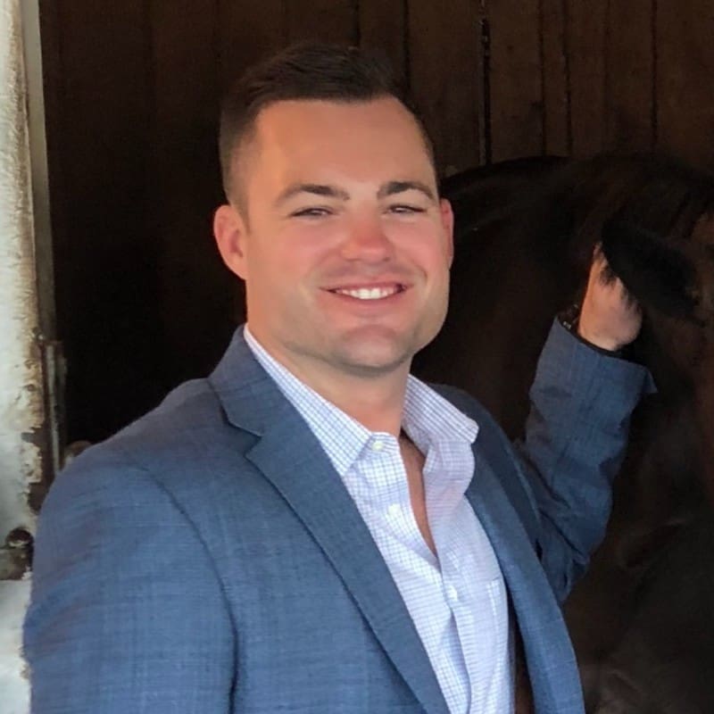A man in a light blue blazer and light-colored shirt smiles at the camera while standing next to a dark horse in a wooden stable.