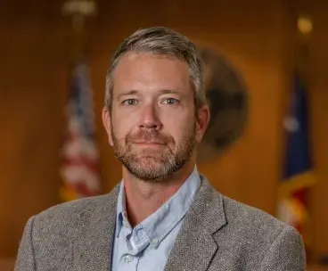 A man with short gray hair and a beard wearing a gray blazer and light blue shirt stands in front of blurred flags and a wooden background.