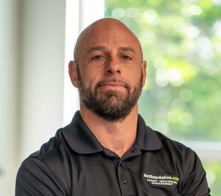 A man with a shaved head and beard wearing a black Netreputation.com polo shirt stands indoors, looking at the camera with a neutral expression. Bright natural light and greenery are visible in the background.