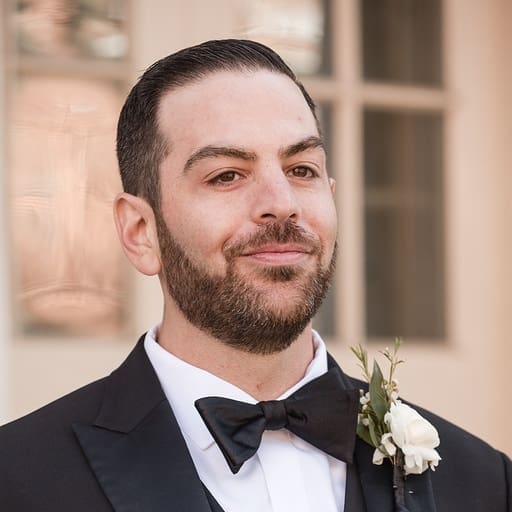 A man with dark hair and a beard, dressed in a black tuxedo with a bow tie and white boutonniere, smiles slightly while standing outdoors in front of a windowed building, ready to celebrate with his team.