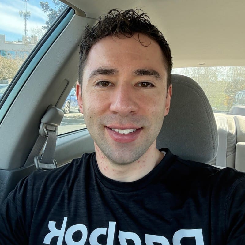 A man with short curly brown hair smiles while sitting in a car, wearing a black Reebok shirt. Sunlight streams through the windows, and the background reveals a parking lot and trees—perhaps he’s just finished a workout with his team.