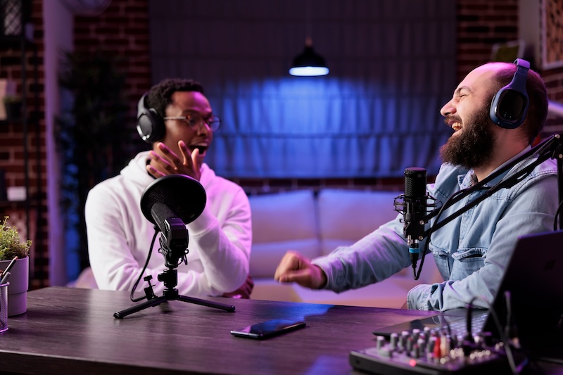 Two men recording a podcast on reputation management for celebrities in a studio with microphones and audio equipment. One man laughs while the other listens intently, both wearing headphones.