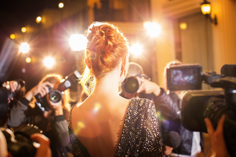 A woman in a sparkling black dress is viewed from behind, surrounded by paparazzi with cameras flashing at a glamorous event focused on reputation management for celebrities.