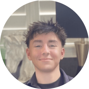 A young team member with short, tousled dark hair smiles at the camera indoors, with white blinds and a lamp in the softly blurred background.