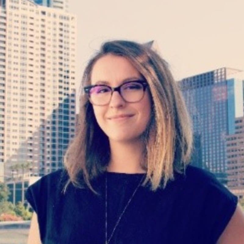 A woman with shoulder-length brown hair and glasses, wearing a black top, stands outdoors in front of tall city buildings on a sunny day.