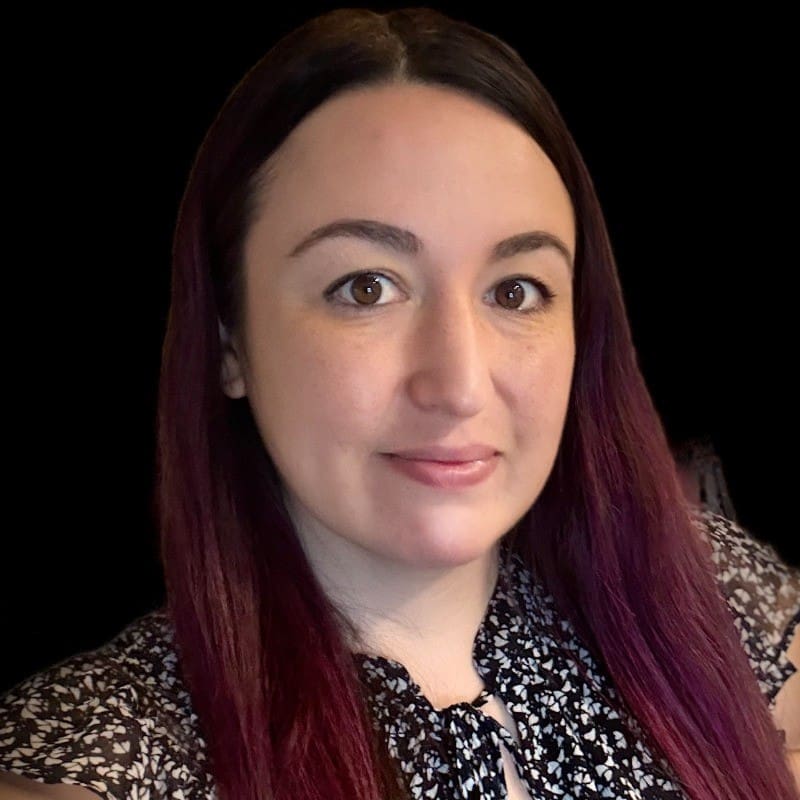 A woman with long, straight, dark hair and fair skin smiles softly at the camera. She is wearing a patterned black and white blouse and is posed against a plain black background.