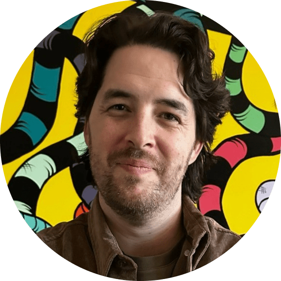 A man with dark, wavy hair and a beard smiles softly. He is wearing a brown jacket, standing before a colorful, abstract background of yellow, black, turquoise, and red patterns—radiating warmth like the heart of a team.