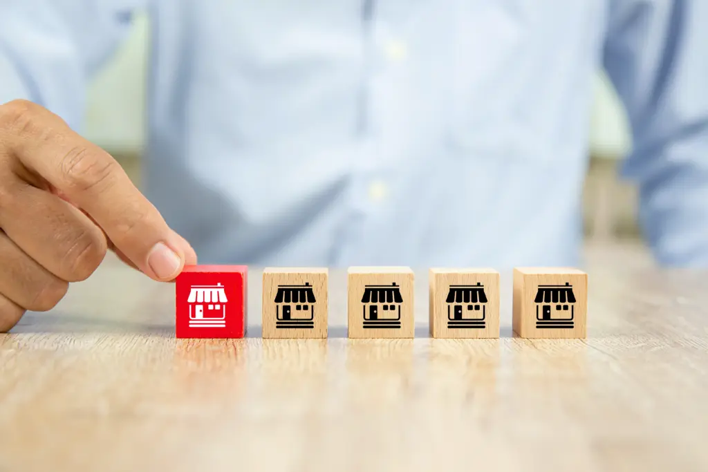 A person’s hand picks up a red wooden block with a white storefront icon from a row of four beige blocks, each with identical black storefront icons, sitting on a wooden table.