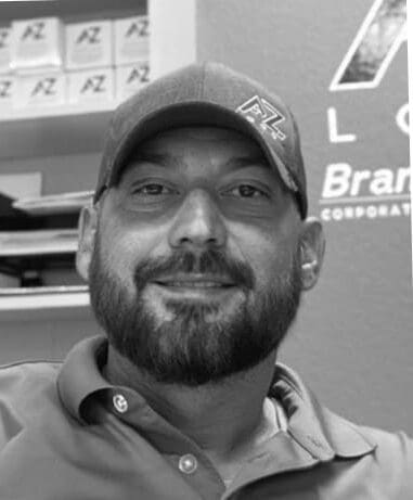 A bearded man wearing a cap and polo shirt smiles at the camera in an office, with shelves and branded signage visible in the background.