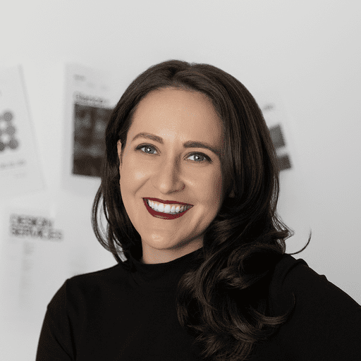 A woman with long dark hair and bright red lipstick smiles at the camera, wearing a black top. Blurred papers or team posters are visible on the white wall behind her.