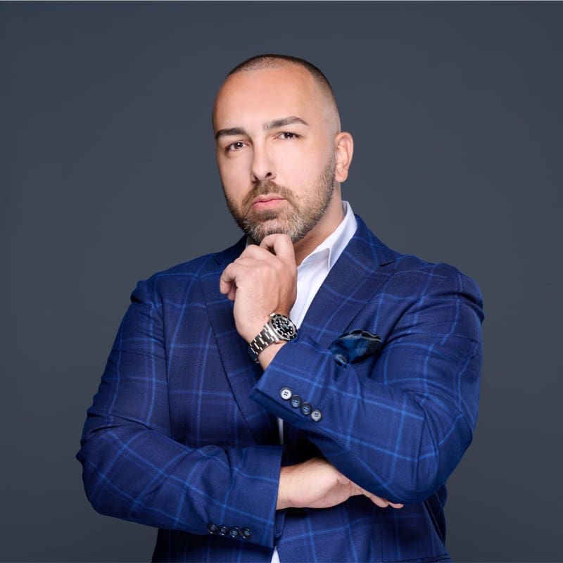 A man with a trimmed beard and shaved head wearing a blue plaid suit jacket and white shirt poses thoughtfully with one hand on his chin against a plain dark background.