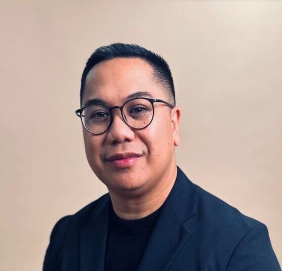 A man with short dark hair, glasses, and a black blazer over a black shirt sits against a plain beige background, facing the camera and smiling slightly.