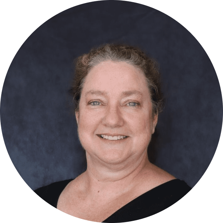 A middle-aged woman with light skin, blue eyes, and curly brown hair pulled back, smiling at the camera against a dark, solid background. She is wearing a black top, radiating a warm and approachable team spirit.