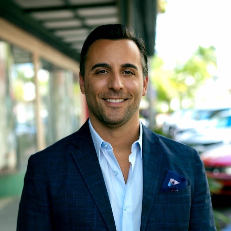 A man in a blue suit jacket and open-collared shirt smiles while standing on a city sidewalk with parked cars and storefronts in the background.