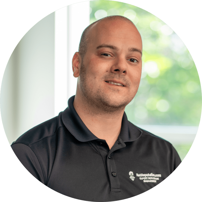 A man with a shaved head wearing a dark collared shirt sits indoors, smiling slightly. The background shows blurred green trees and sunlight through a window.