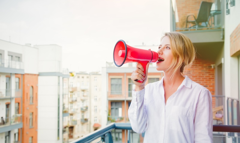 A woman on a balcony is speaking into a red megaphone, surrounded by apartment buildings during a sunny day, advocating for online reputation management.