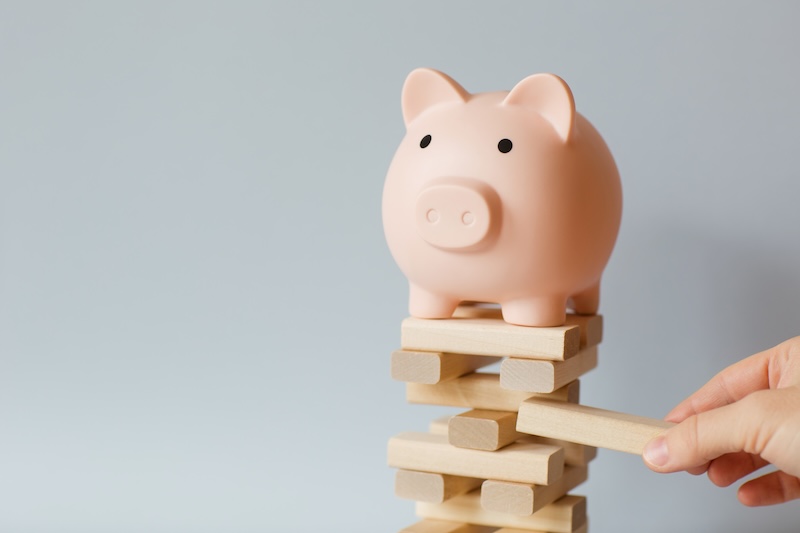 A pink piggy bank stands on top of a precarious stack of wooden blocks, representing Fortune's Most Admired Companies. A human hand is adding another block to the unstable structure, symbolizing growth