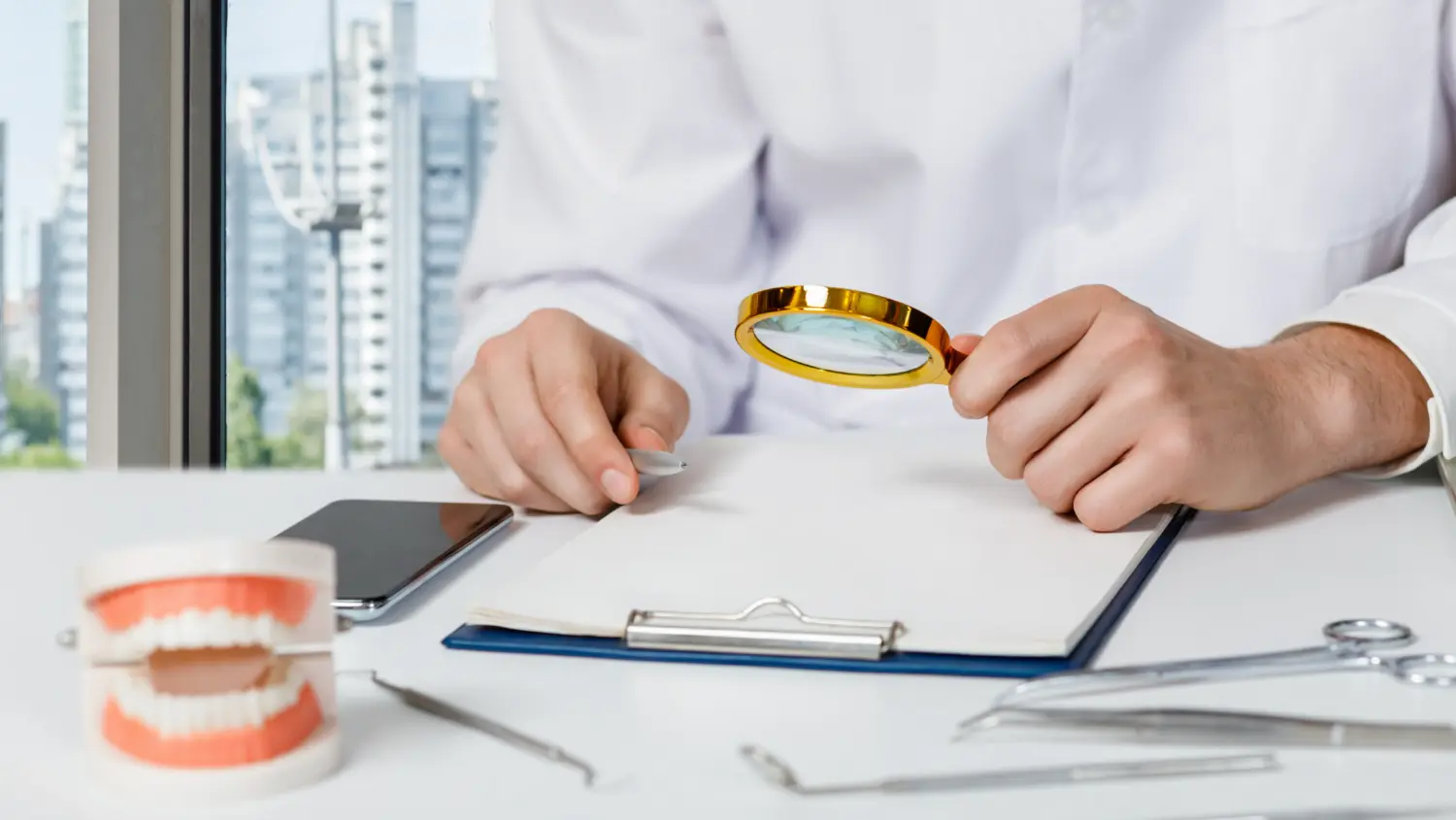 A person in a white coat holds a magnifying glass over a clipboard on a desk with dental tools, a model of teeth, and a smartphone, with a cityscape visible through the window.