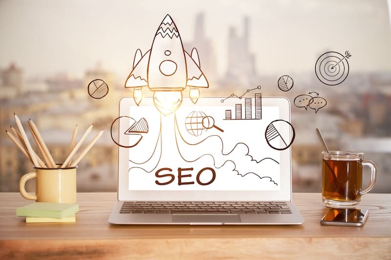 A laptop on a desk displays the word "SEO" with a rocket and business-related doodles, including graphs and target icons. Nearby are pencils in a cup, a glass of tea, and a cityscape in the blurred background.