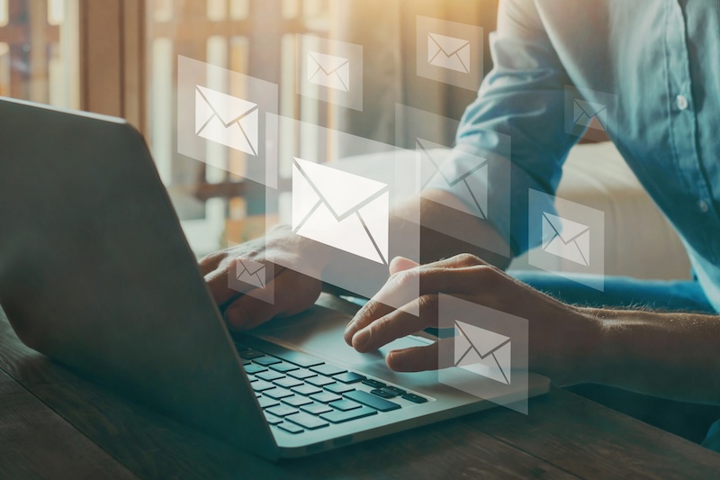 A person typing on a laptop at a desk, with illustrated email icons floating above the keyboard, representing sending or receiving emails in a digital work environment or searching "how do I remove my name from Google.