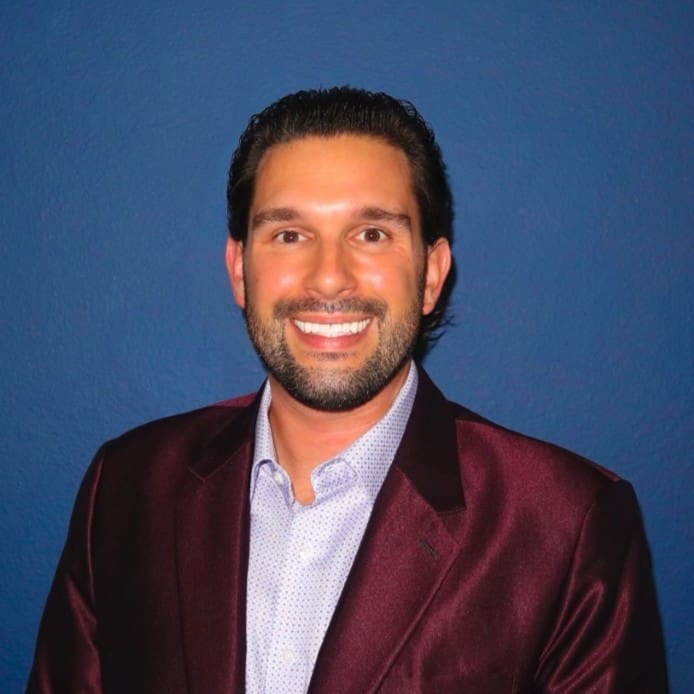 A man with short dark hair and a beard, wearing a maroon blazer and light-colored shirt, smiling confidently in front of a solid blue background, ready to lead his team.