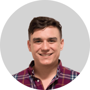 A young man with short brown hair wearing a red and blue plaid shirt smiles at the camera, radiating team spirit against a plain light gray background.