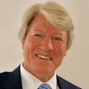 A smiling man with light blond hair wearing a dark suit, white shirt, and blue patterned tie stands in front of a plain light-colored wall.
