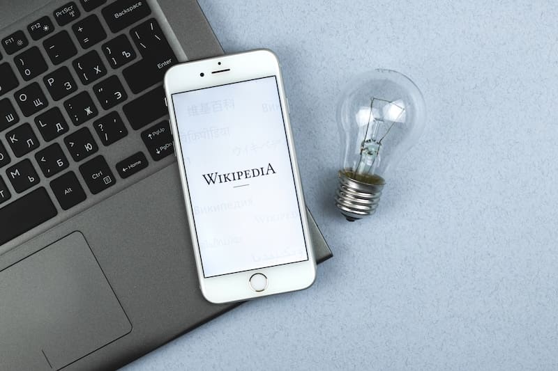 A laptop with the Wikipedia logo on the screen sits on a desk. A phone rests on a laptop. The phone screens shows the Wikipedia homepage. A lightbulb rests to the side.